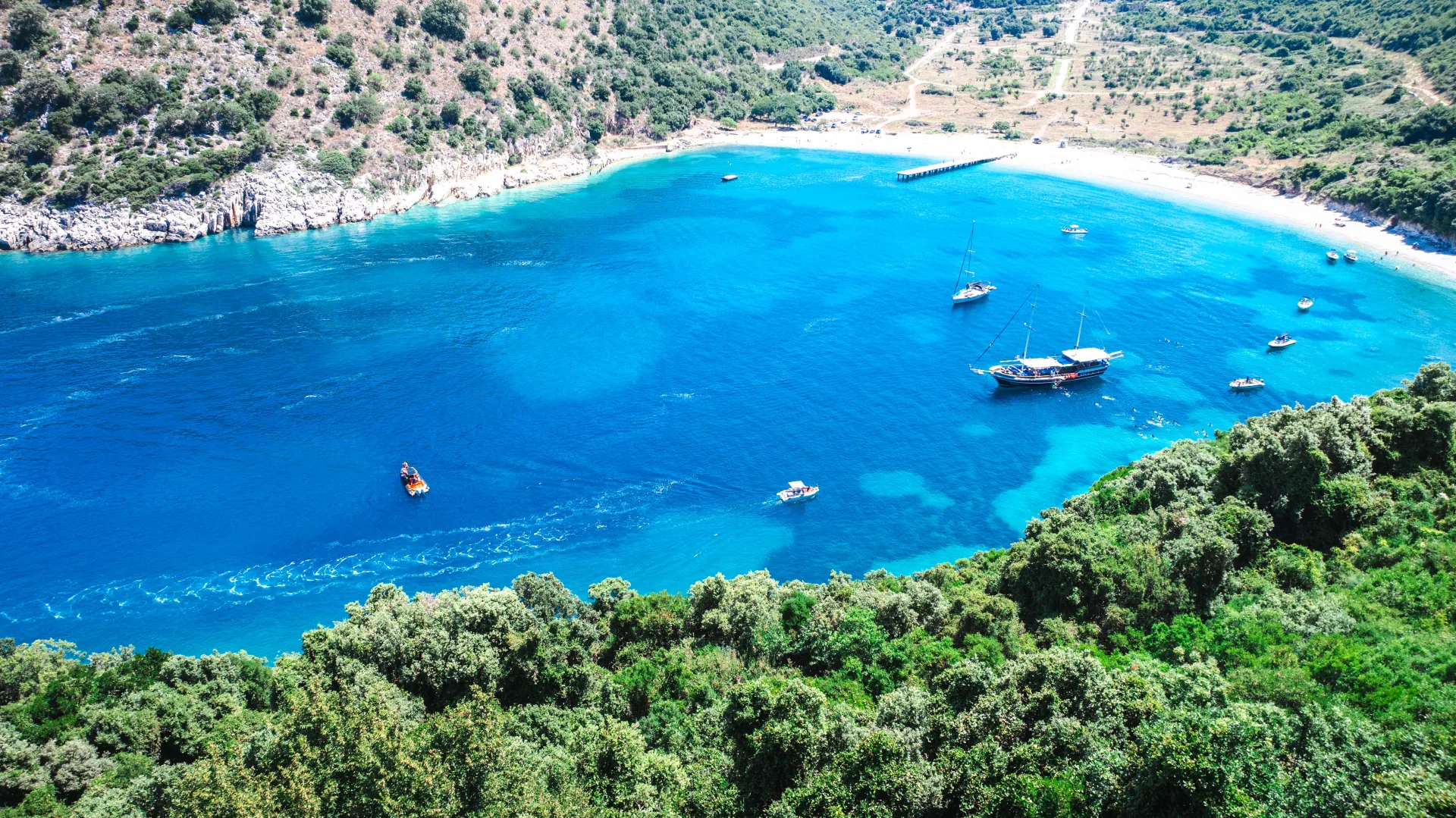 Aerial of a hidden Albanian Riviera cove with a traditional sailing boat and white-sand beach